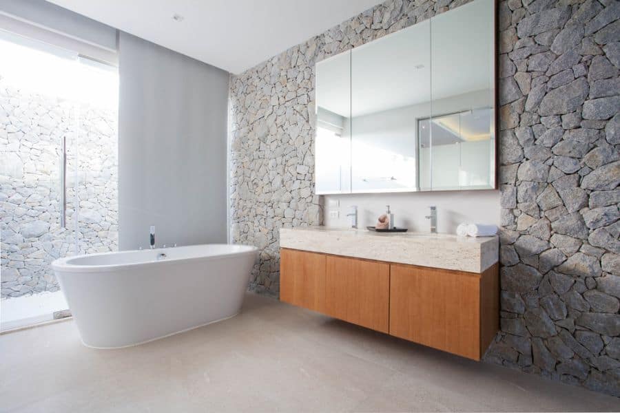 Modern bathroom with a stone accent wall, freestanding bathtub, and wooden vanity topped by a large mirror