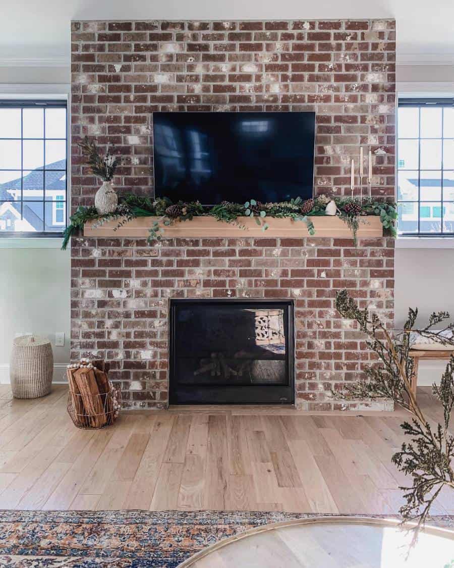 Brick fireplace with TV above and greenery on the mantel, framed by an accent wall and large windows on each side