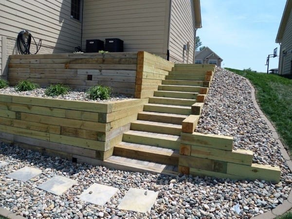 Wooden retaining wall with built-in stairs, surrounded by gravel landscaping on a sloped yard.