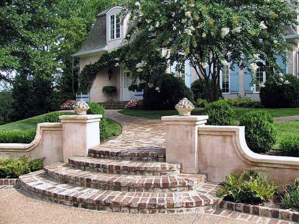 Brick steps and a walkway lead to a house with trees and shrubs in the front yard