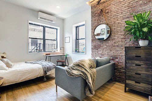 Studio with exposed brick wall, cozy bed, modern sofa, and natural light from large windows.