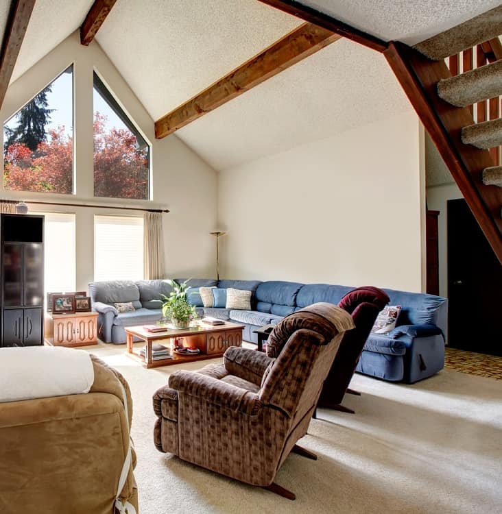 A bright and spacious living room featuring a vaulted ceiling with wooden beams, large windows offering natural light, and comfortable seating for a relaxed atmosphere