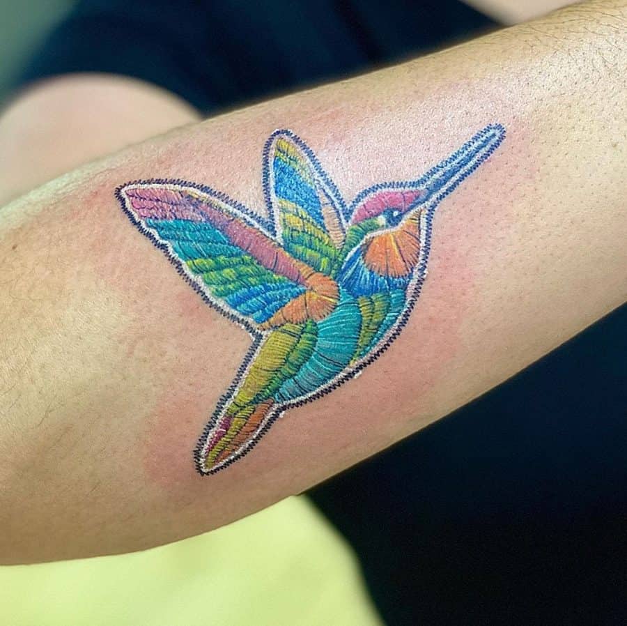 Vibrant tattoo of a hummingbird in flight, designed with an embroidery effect that mimics fabric stitching.