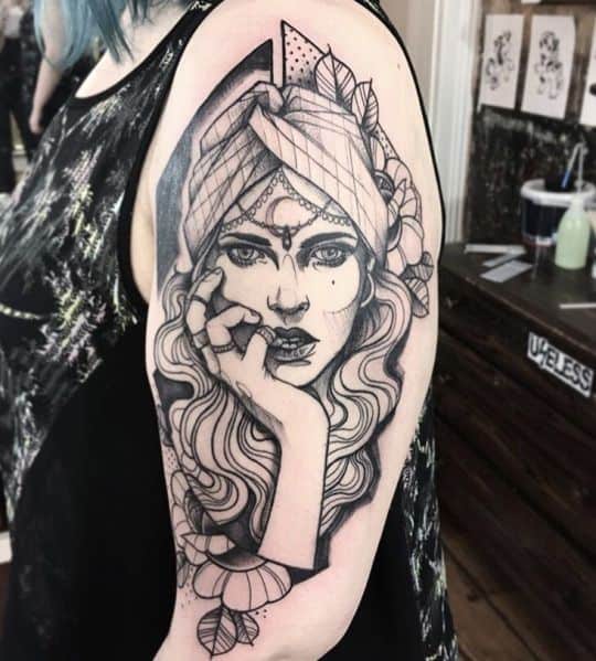 Black and grey tattoo of a mystical gypsy woman with piercing eyes, an intricate headscarf, and floral details on the upper arm.