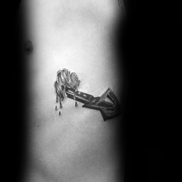 Black and grey tattoo of a broadhead arrow piercing through skin with realistic blood drops and torn flesh details.