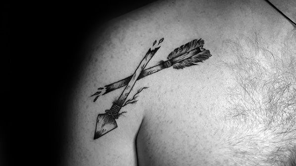 Black and grey tattoo of a broken arrow crossing itself on the shoulder, detailed with cracks, wood texture, and feather shading.