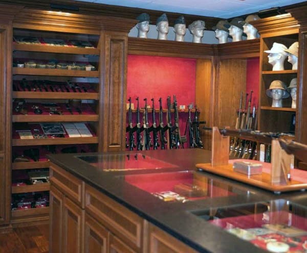 Gun room with wooden cabinetry, display cases, rifles, and shelves of collectible items.