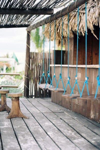 Rustic tiki bar with hanging swing seats and wooden stools on a deck with thatched roof.