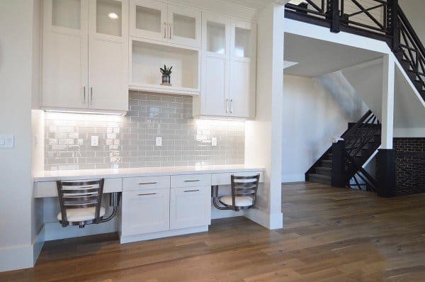 Modern home office area with a built-in desk, two chairs, white cabinets, and tiled backsplash by the staircase