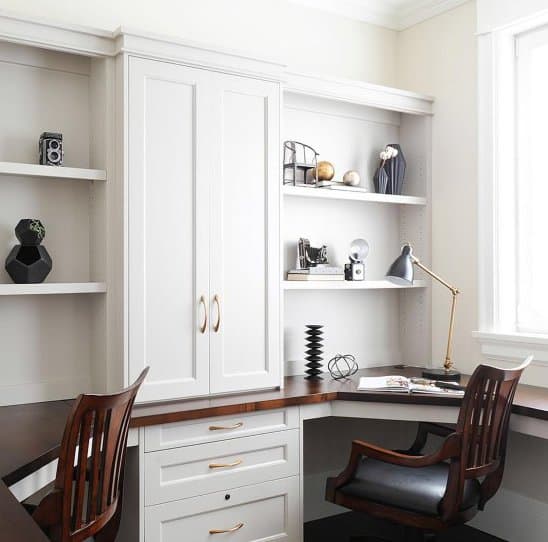 A corner office with white shelves, built-in desk, chairs, and decorative items, lit by natural light from a window
