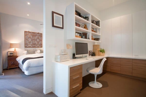 Modern home office with built-in desk, chair, shelves, and computer; adjacent to a cozy bedroom with a neatly made bed