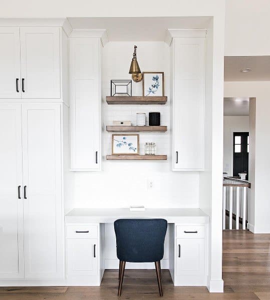 Built-in minimalist white desk nook with wooden shelves, a black chair, and decorative items in a bright room