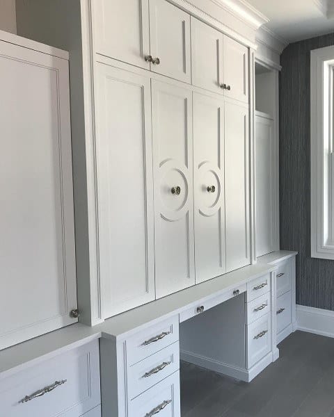 White built-in cabinets, drawers, and desk with modern handles, next to a large window