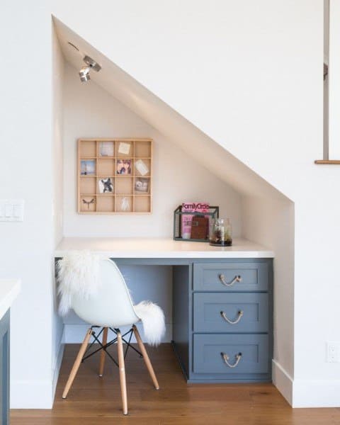 Cozy home office nook with a built-in desk, white chair, blue drawers, wall shelf, and decor under a slanted ceiling