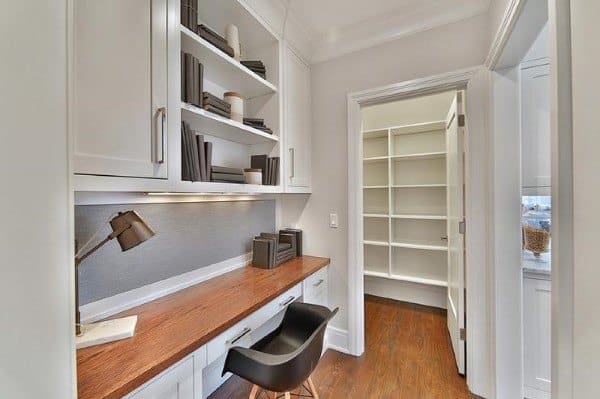 Home office with a built-in wooden desk, shelves, black chair, and an open door to a storage room with empty shelves