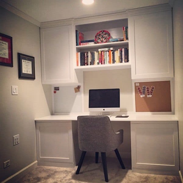 Home office with built-in desk, computer, chair, bookshelf, and corkboard; framed certificates adorn the walls