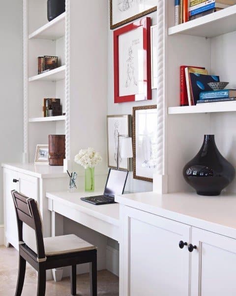 A white home office with a built-in desk, shelves, books, a black vase, framed art, and flowers