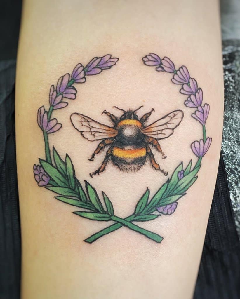 A tattoo of a bumblebee surrounded by a wreath of lavender and green leaves, with the stems forming a cross beneath the bee.