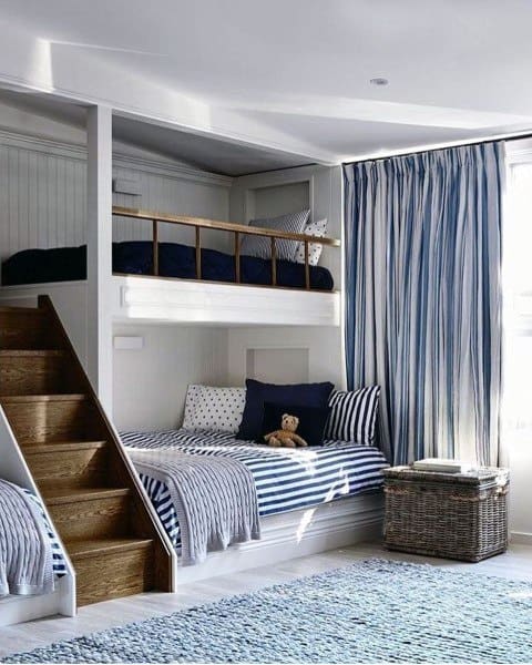 Cozy bedroom with bunk beds, striped bedding, blue curtains, and wooden staircase.