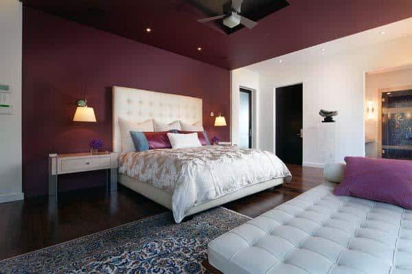 Modern bedroom with a beige bed, red accent wall, ceiling fan, and a white chaise lounge on a patterned rug