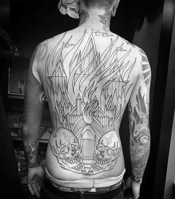 Large back tattoo of a burning cathedral with towering flames and gothic architecture. Two mirrored skulls appear at the lower back.