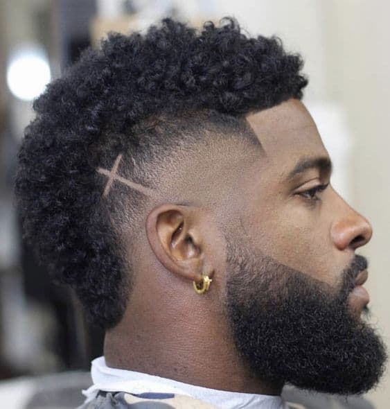 A burst fade haircut styled on curly hair and a cross design created on one side. A clean cut hairline continues to the beard