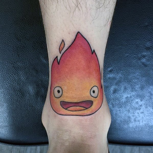 Minimalist Calcifer tattoo with a bright flame shape, wide eyes, and a cheerful open-mouth expression on the back of the leg.