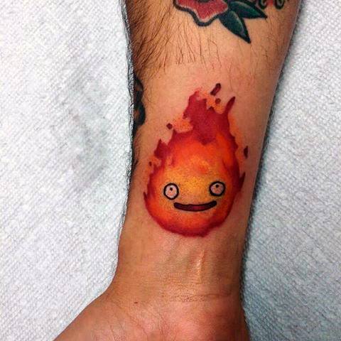 Small Calcifer tattoo on the wrist with a calm smile, glowing orange-red flames, and a soft, friendly look.