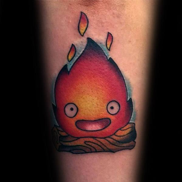 Bold Calcifer tattoo with vibrant orange and red flames, perched on a log, smiling with wide eyes and small flickers above his head.