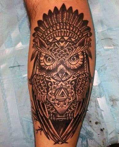 Intricate black and grey calf tattoo of an owl with geometric patterns and tribal-inspired details, featuring intense, piercing eyes