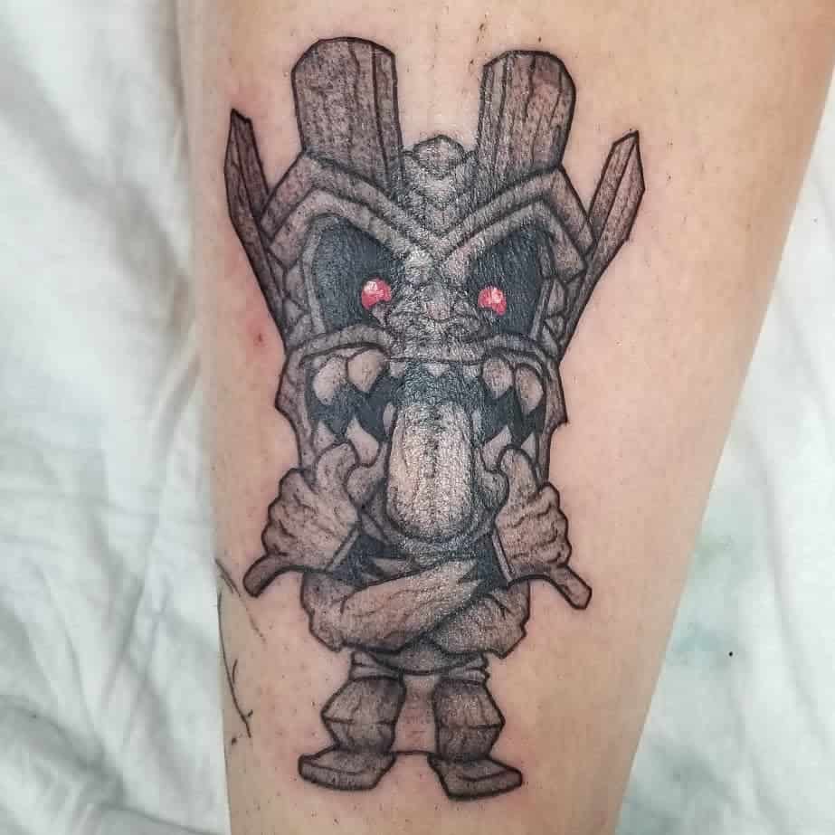 Black and grey Tiki tattoo featuring a cartoonish mask with a protruding tongue, red eyes, sharp teeth, and a playful pose