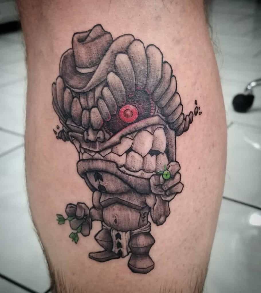 Black and grey Tiki tattoo featuring a cartoonish mask with a cowboy hat, sharp teeth, and green darts and green ball in its hands