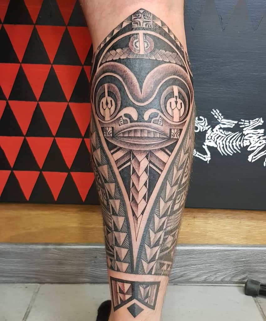 Intricate black and grey Tiki tattoo on the leg, featuring a detailed mask with geometric patterns and sharp lines