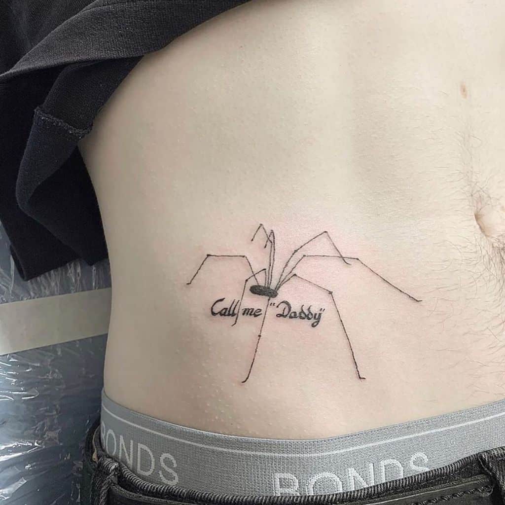 A fine-line tattoo of a daddy longlegs spider with the phrase 'Call me Daddy' integrated into its body, placed on the ribcage.