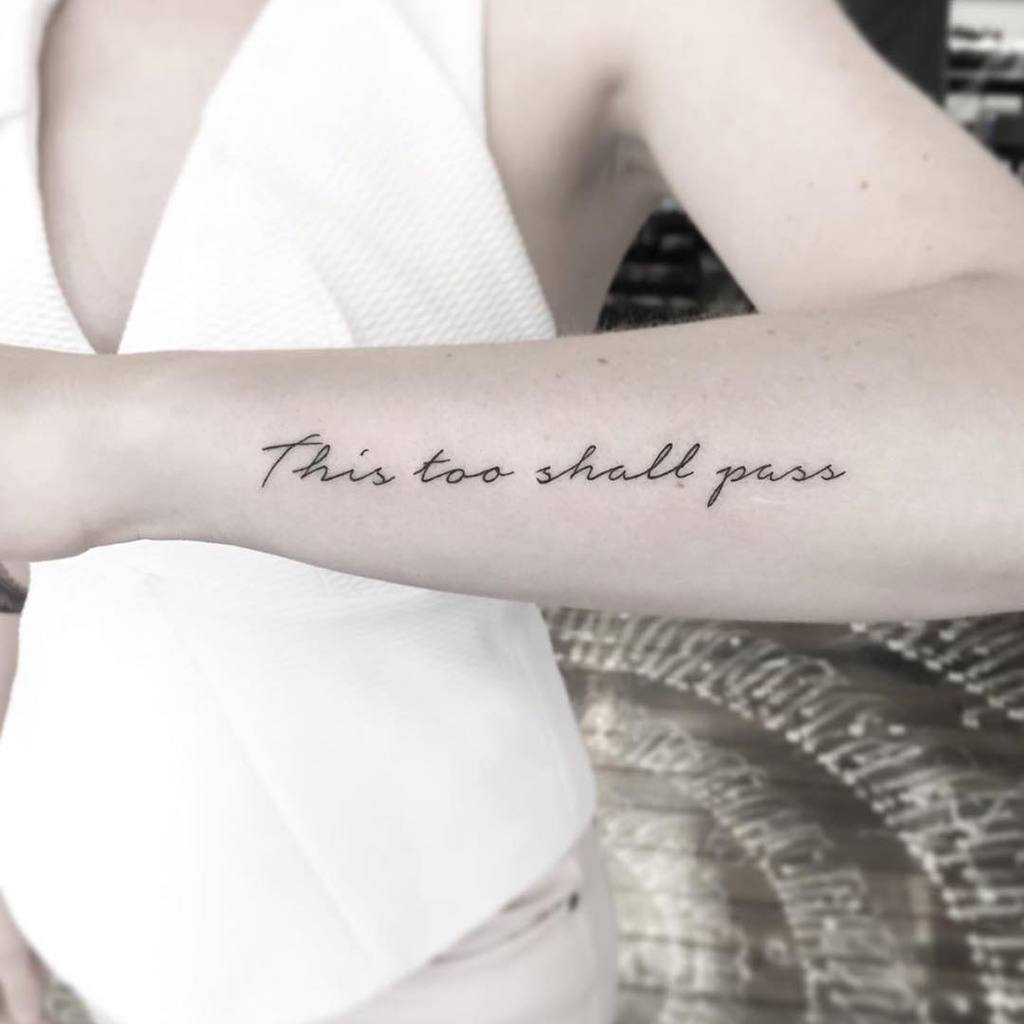 A "This Too Shall Pass" tattoo in cursive on a person's forearm, with a minimalist white top visible in the frame.
