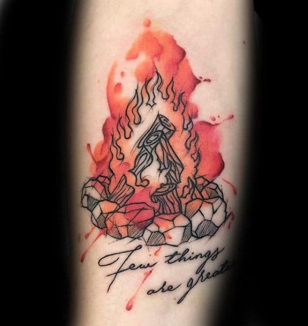 Campfire tattoo with watercolor-style red flames, outlined logs and stones, and cursive text reading “Few things are greater”.