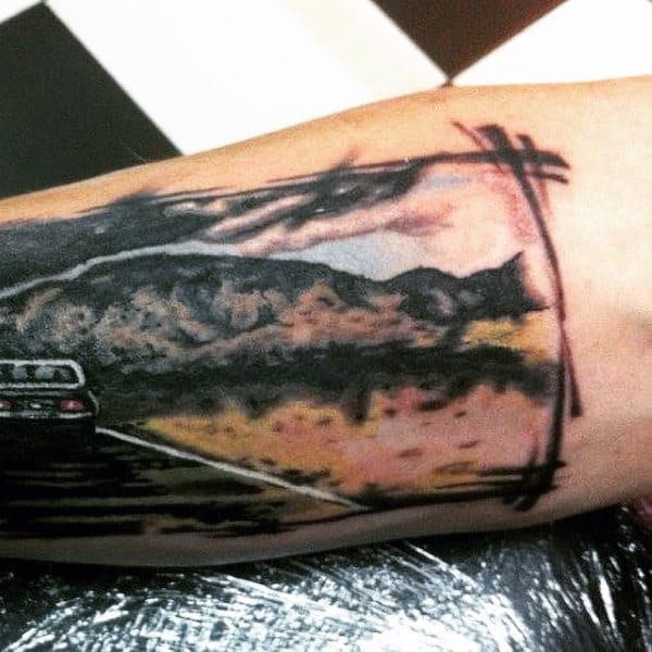 Forearm tattoo of a car on a road heading towards distant mountains under a framed black and white background