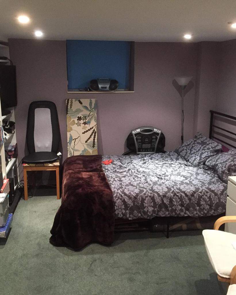 Small basement bedroom with purple walls, patterned bedding, dark wood bedframe, and a compact shelving unit.
