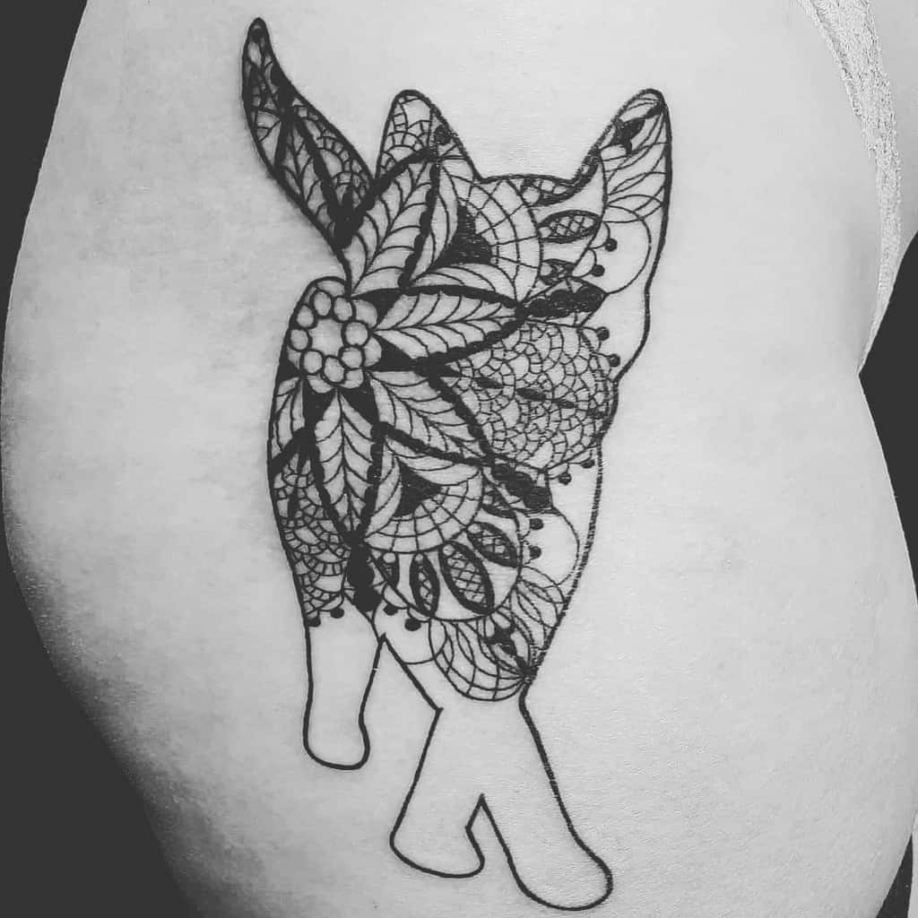 Tattoo of a cat walking away, with intricate lace and floral patterns filling one half of its body, blending realism with ornamental design.