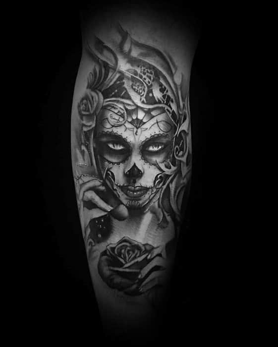 La Catrina tattoo of a woman with haunting eyes, sugar skull makeup, ornate headpiece, and a rose held near her lips in black and grey.