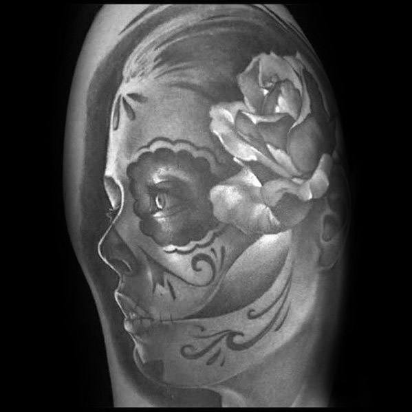 La Catrina tattoo of a woman in profile with sugar skull makeup and a large, realistic rose over her ear in soft black and grey tones.