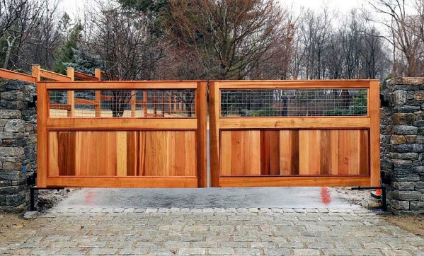 Cedar wood driveway gate with wire mesh panels framed by stone columns.
