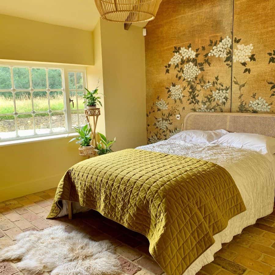 Cozy bedroom with a floral accent wall, large window, and a yellow quilted bed. Elegant decor with plants adds charm