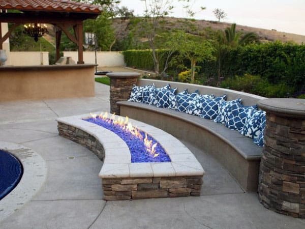 Curved outdoor stone bench with blue pillows and a long fire pit, surrounded by greenery and a hill backdrop