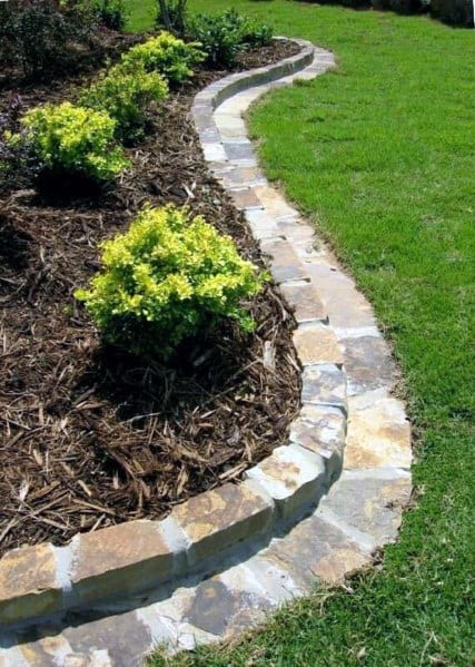 A curved stone border around a garden bed with lush green plants and mulch, adding structure to the lawn