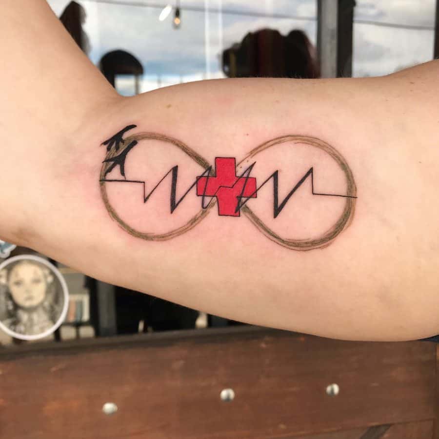 This tattoo features an infinity symbol with a heartbeat line crossing through it, paired with a red medical cross, and a crow flying in the design. The combination evokes themes of life, eternity, and healing.