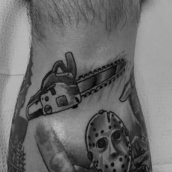 Black and gray horror tattoo featuring a detailed chainsaw above a menacing masked figure, blending slasher and thriller elements.
