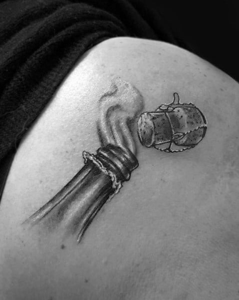 Tattoo of a smoking bottle neck with a cork flying off, inked on a person's shoulder.