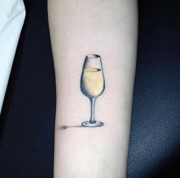 Tattoo of a champagne glass on an arm, with realistic shadow detail, against a black background.