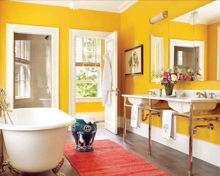 A bright yellow bathroom with a white tub, red rug, double sinks, and a blue garden stool providing a playful accent.